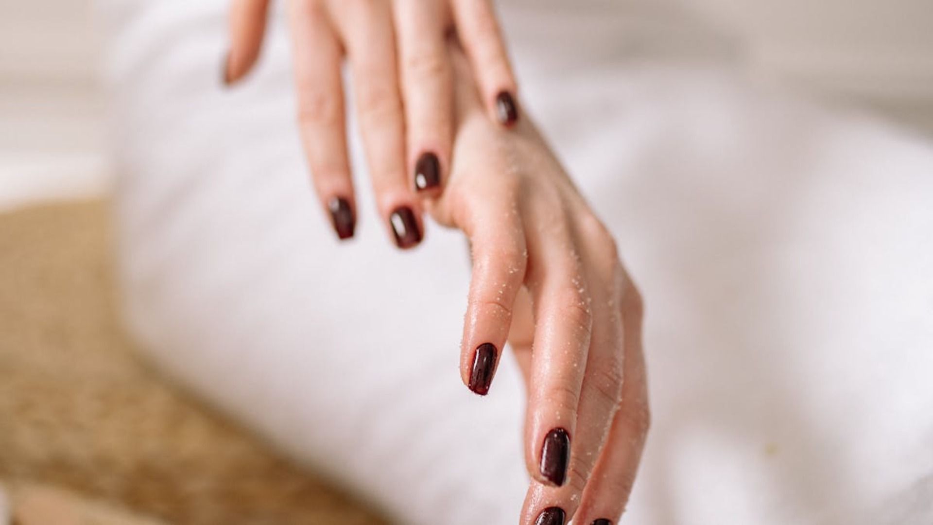 Manos con esmalte de uñas color burdeos oscuro, apoyadas sobre tela blanca.