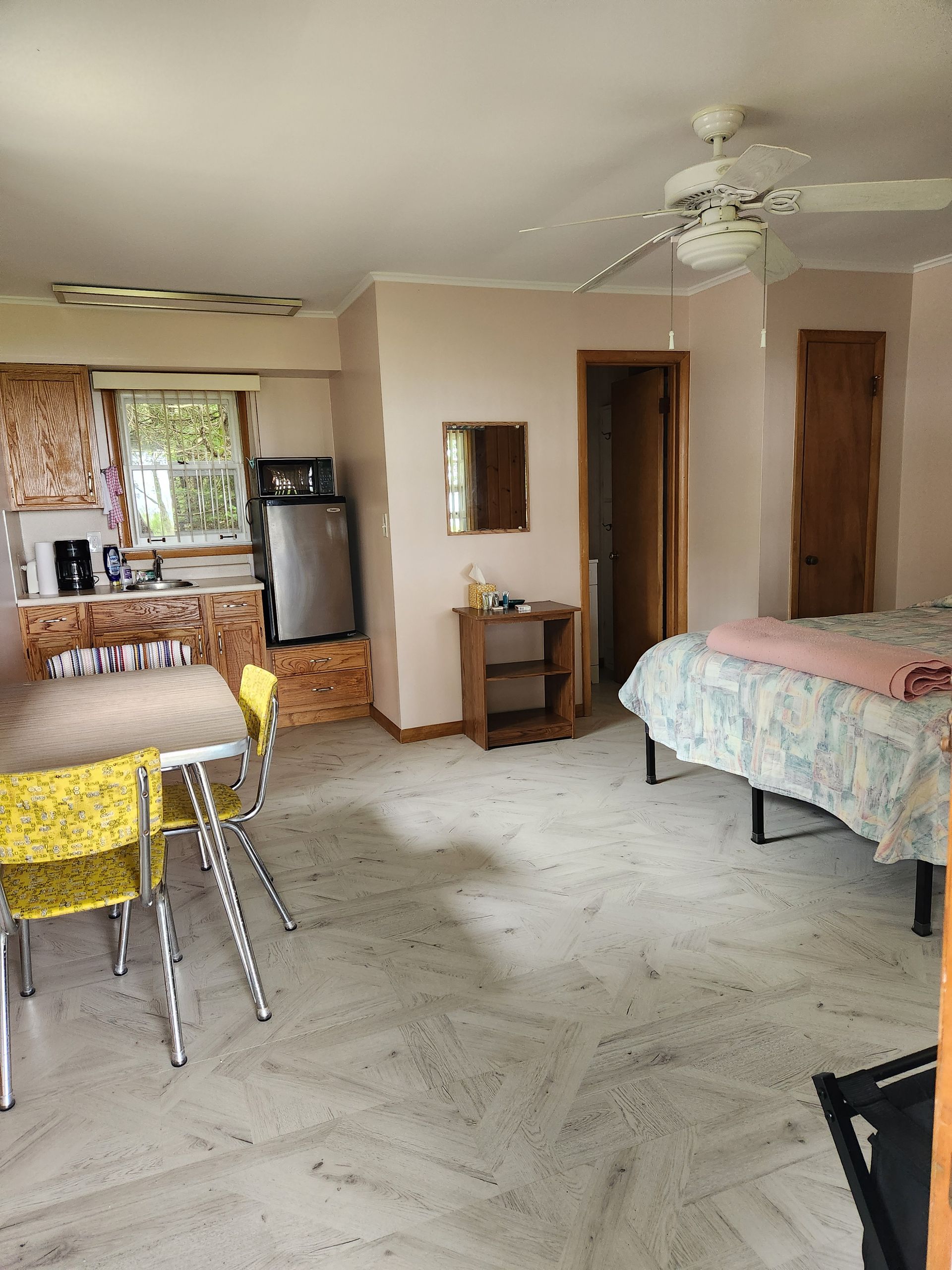 Small studio apartment with a bed, kitchen, table, and yellow chairs. Pink walls and a ceiling fan.