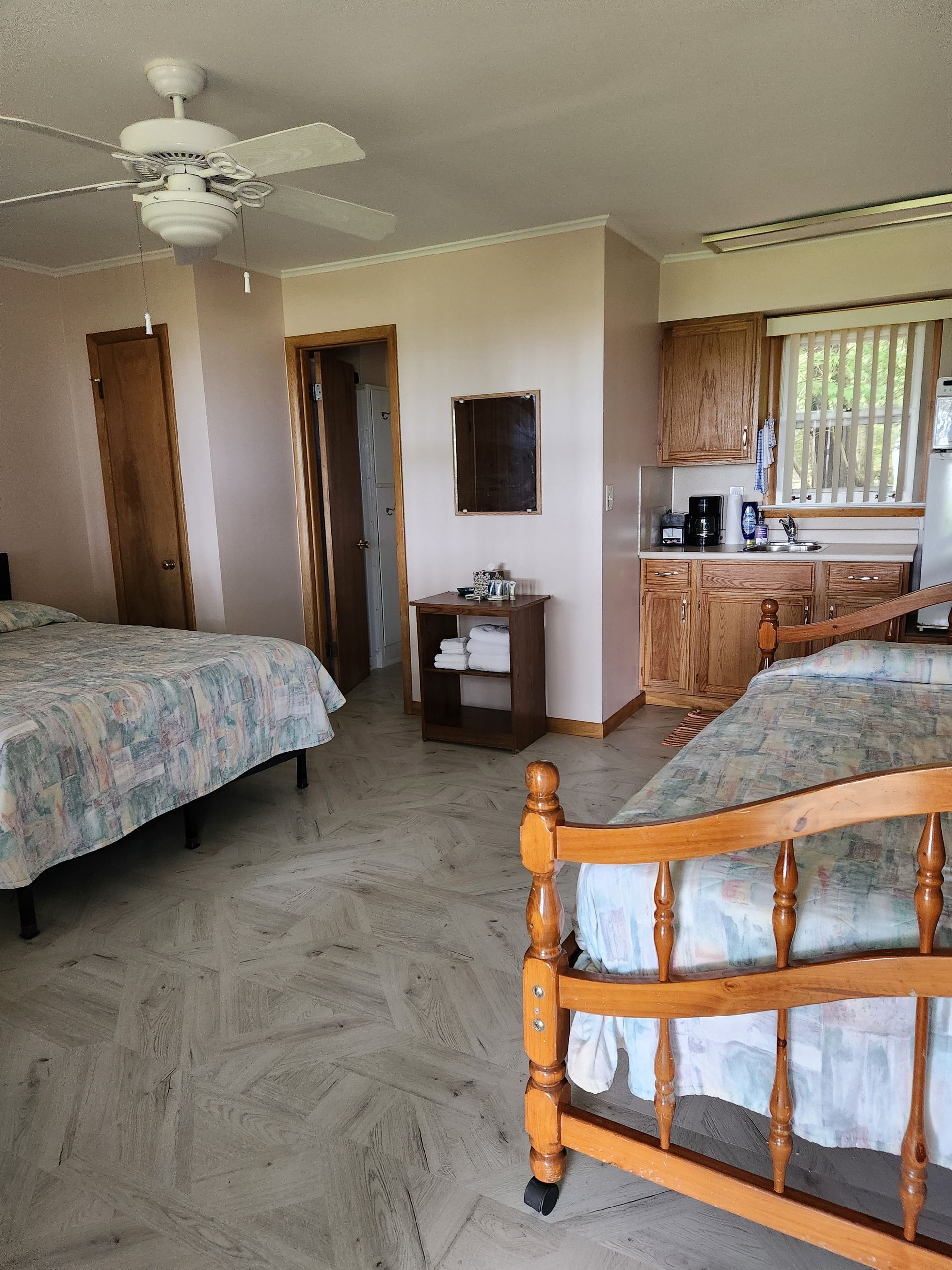 Bedroom with two beds, kitchenette, light-colored walls, ceiling fan, and wooden furniture.