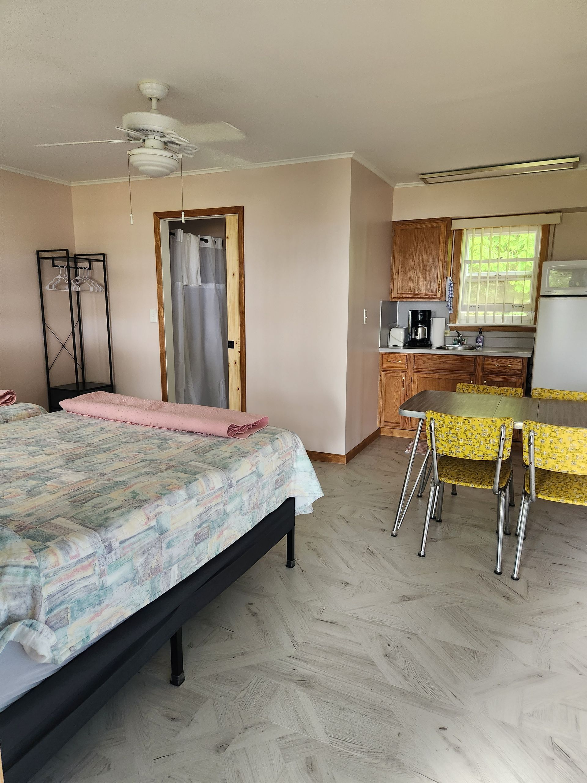 Bedroom with bed, small kitchen, and table. Light-colored walls, patterned bedspread, and concrete floor.