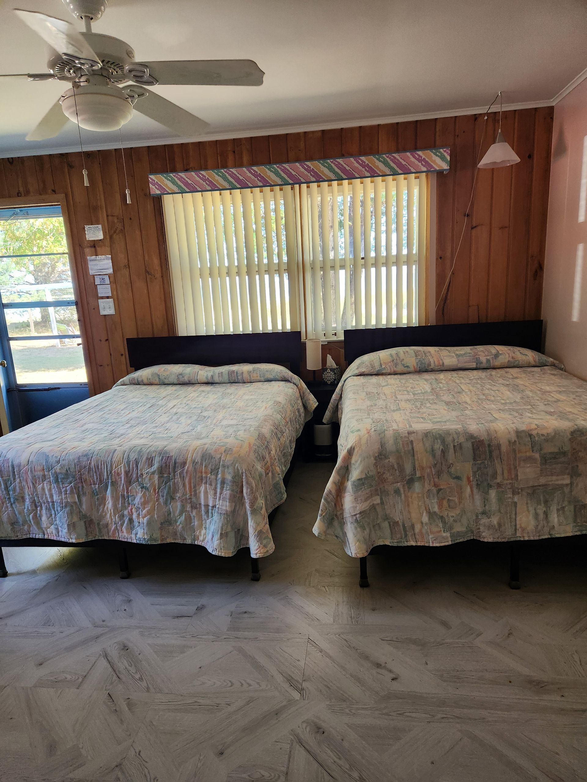 Two beds in a room with wood paneling, a window with blinds, and a ceiling fan.