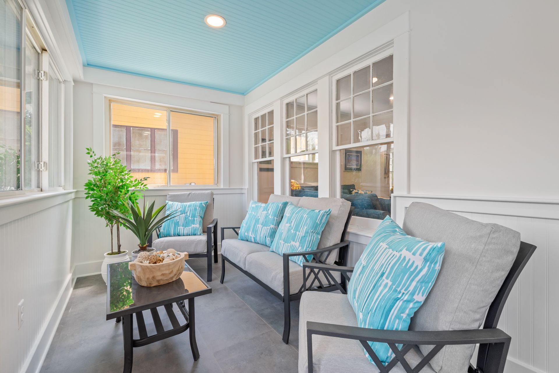 Sunroom with light blue ceiling, white walls, and gray furniture with turquoise pillows.