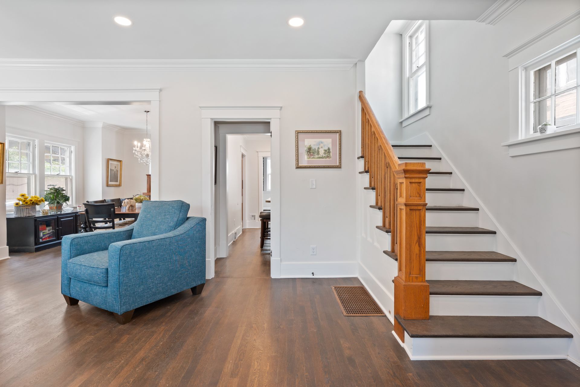 A bright interior with a wooden staircase, teal armchair, and doorway leading to other rooms.