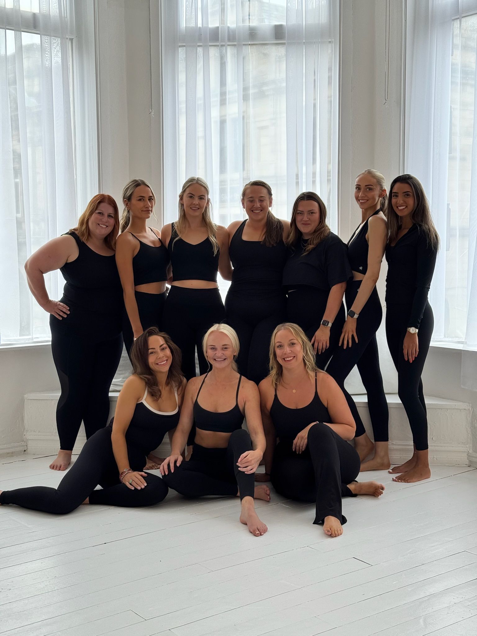 Group of eleven women in black workout clothes posing in a bright room with large windows. Some are standing, others kneeling.