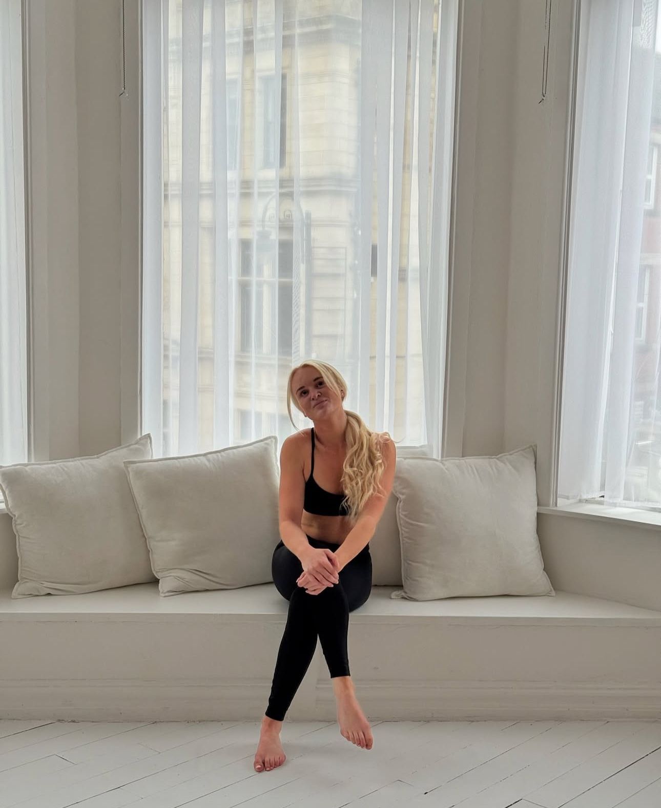 Woman in athletic wear sitting on a white window seat, smiling. She has blonde hair and is in a bright room.