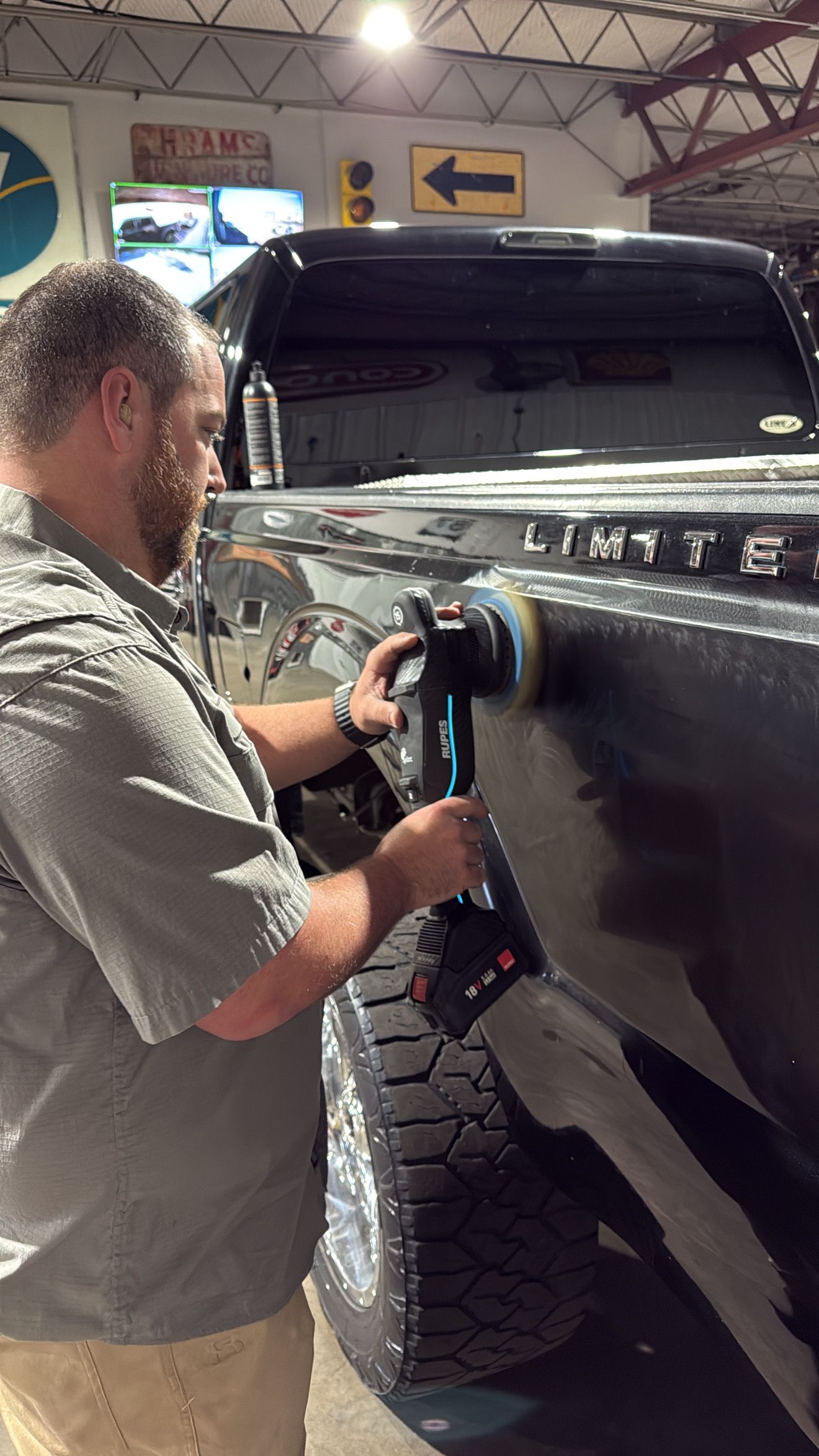 A person uses a power polisher on the side of a black truck inside a garage.