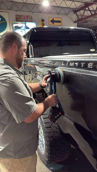 A person uses an electric buffer to polish the black side panel of a large pickup truck in a workshop.