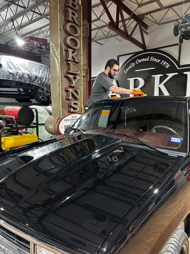 A person wearing orange gloves cleans the windshield of a shiny black truck inside a garage with industrial signage.