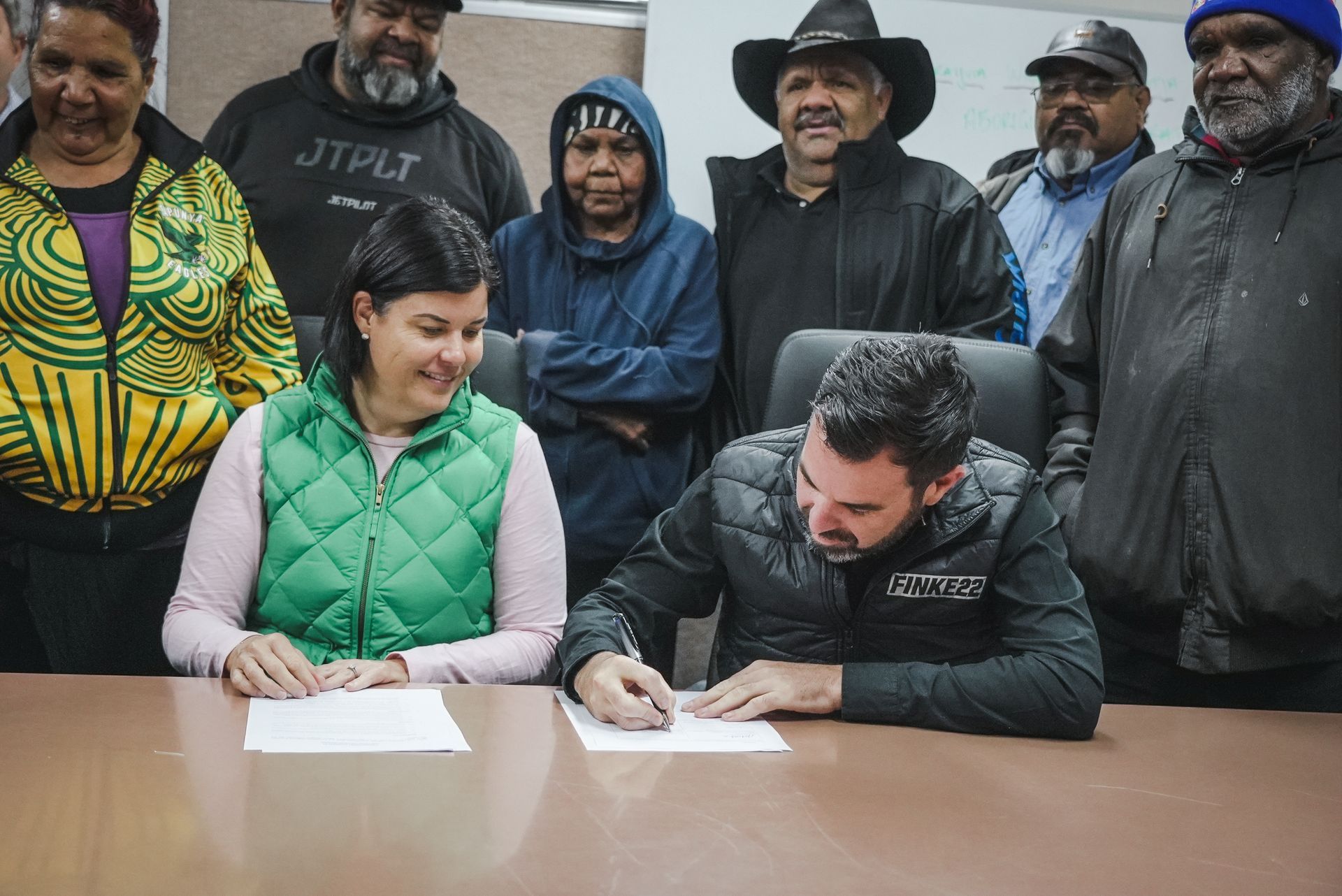 Two people sign papers at a table, watched by a group of people in a room.