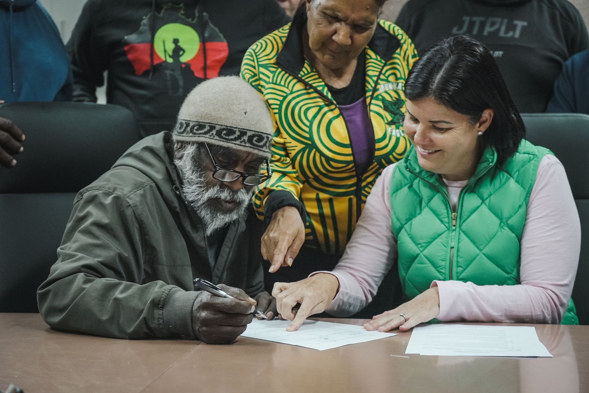 Man signs paper, assisted by a woman, with another woman looking on.
