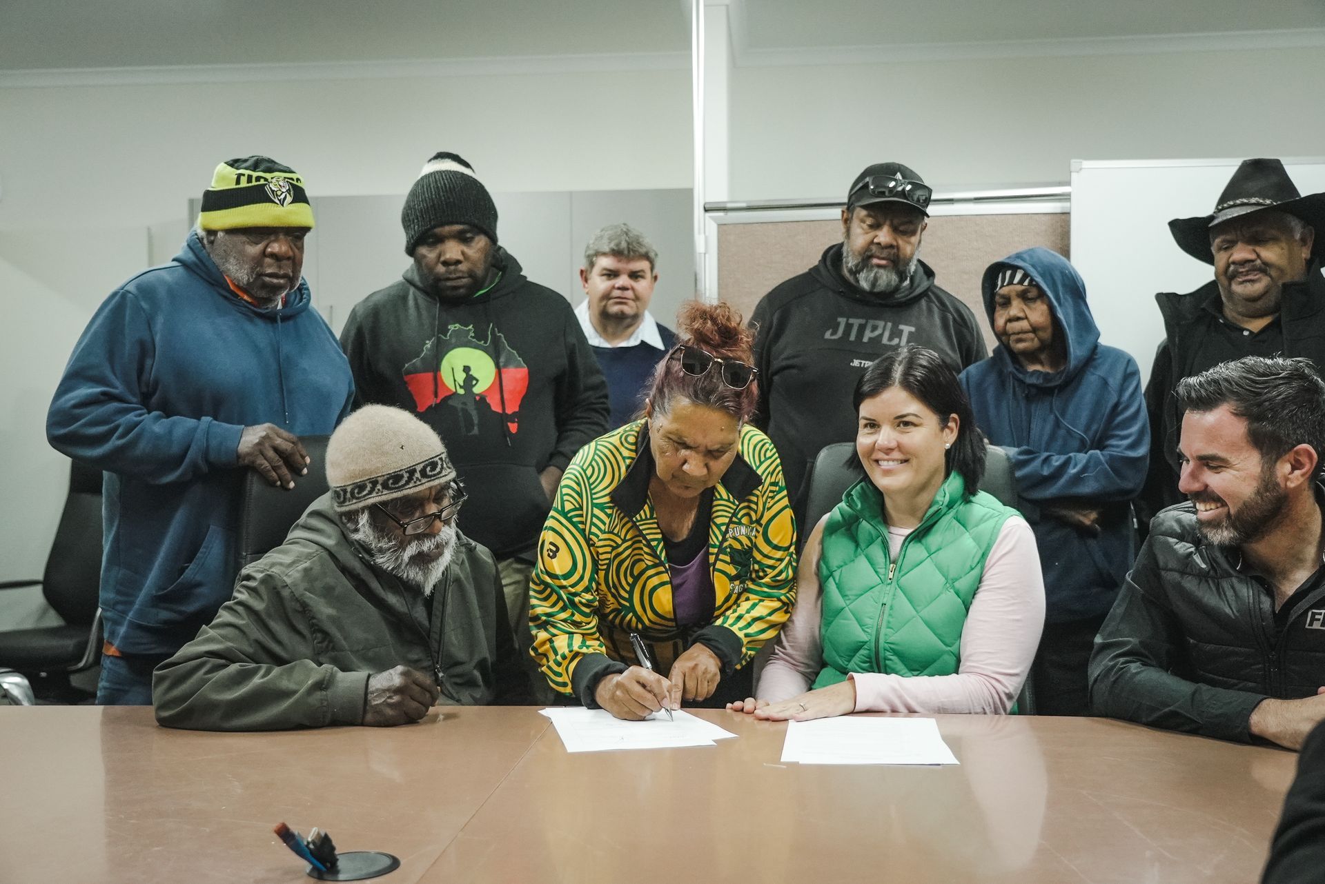 People sign documents at a table. Group surrounds them. Indoors, some wearing hats, smiling.
