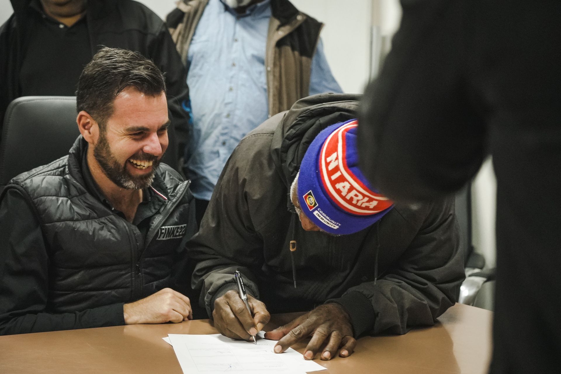 Man signing a document at a table, smiling man watches, others in background.