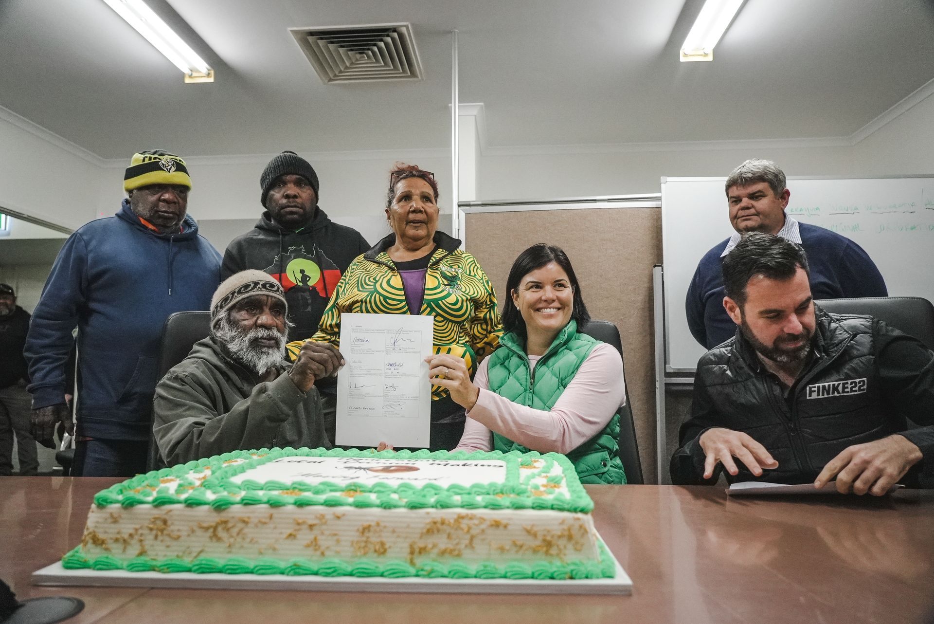 Group of people signing document, cake in foreground, office setting.