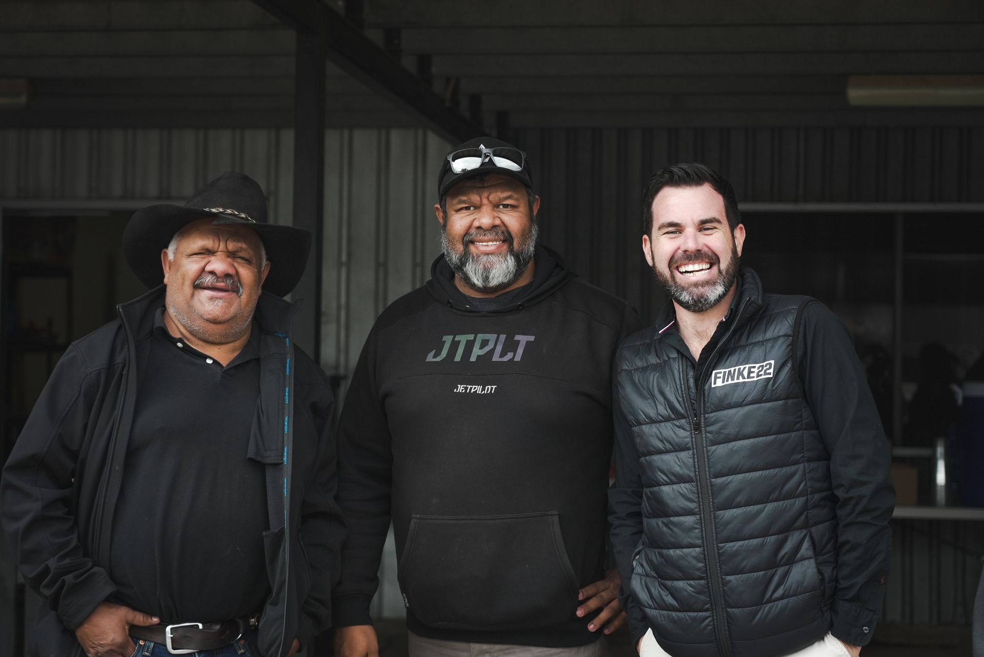 Three men smiling; one in a cowboy hat, one in a hoodie, and one in a vest, in an outdoor setting.
