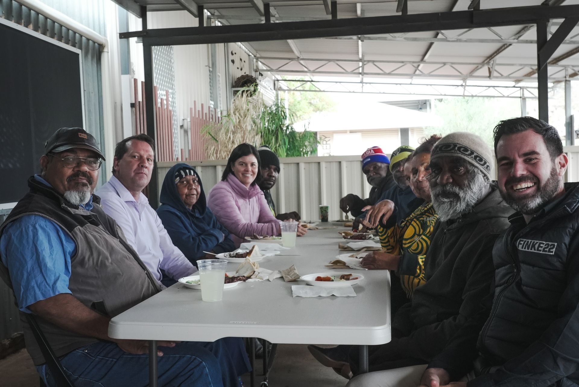 Group of people seated at a table outdoors, smiling. They are eating, under a shaded structure.