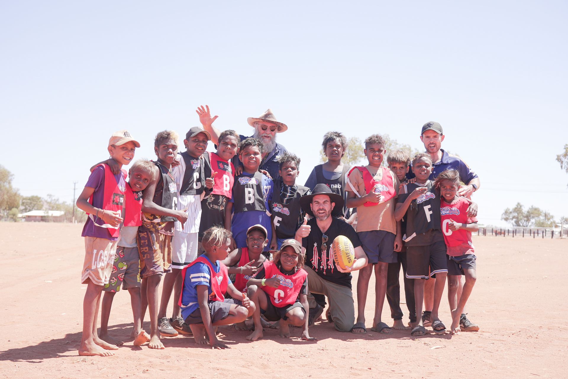 Group of children and adults on a dirt field, smiling, with a football.