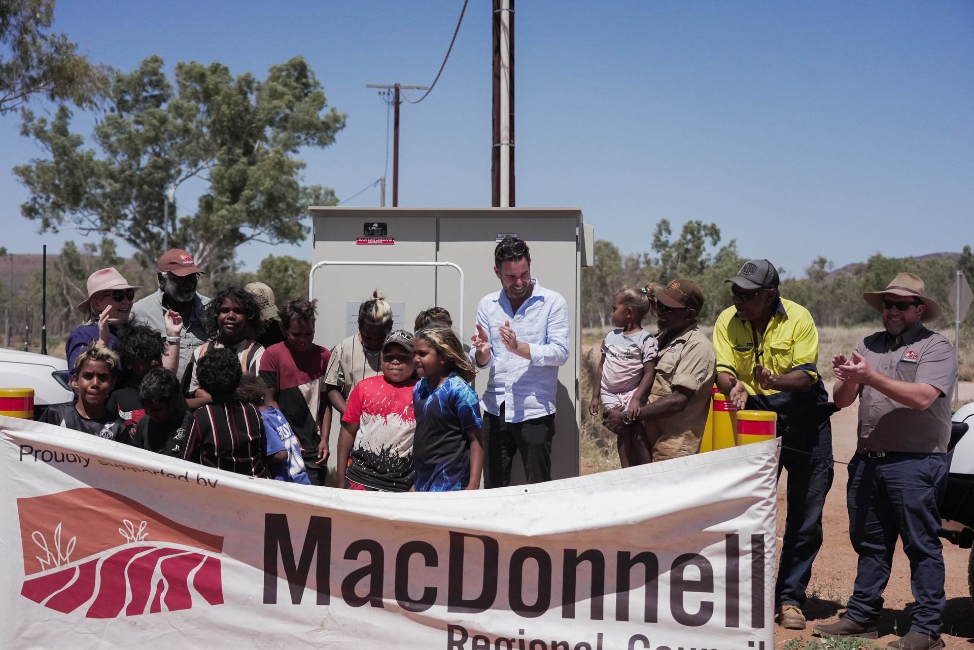People, including children, gather for a ribbon-cutting ceremony near an electrical box.