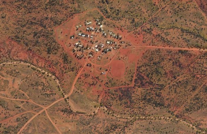 Aerial view of a small cluster of buildings in a reddish-brown desert landscape, with dirt roads leading in and out.