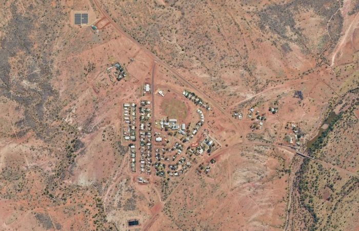 Aerial view of a small desert town with buildings, dirt roads, and surrounding red-brown landscape.