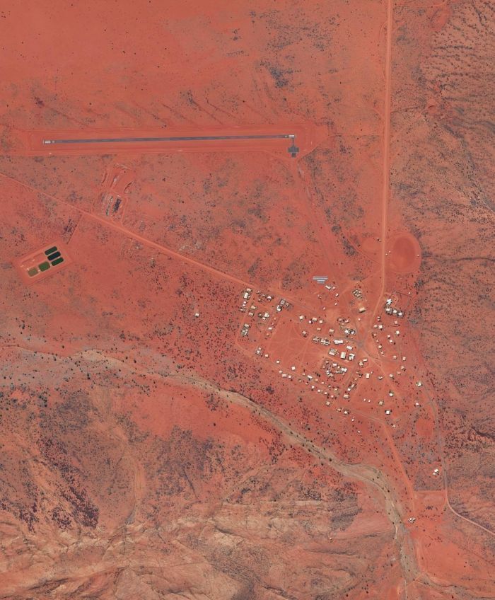Aerial view of a remote settlement with a red dirt airstrip and scattered buildings in a desert landscape.
