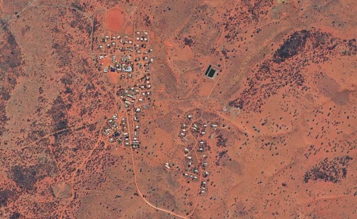 Aerial view of a small settlement in a reddish-brown arid landscape, buildings and scattered trees visible.