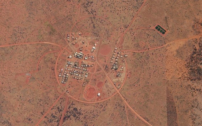 Aerial view of a small village with white buildings, circular roads, and reddish-brown surroundings.