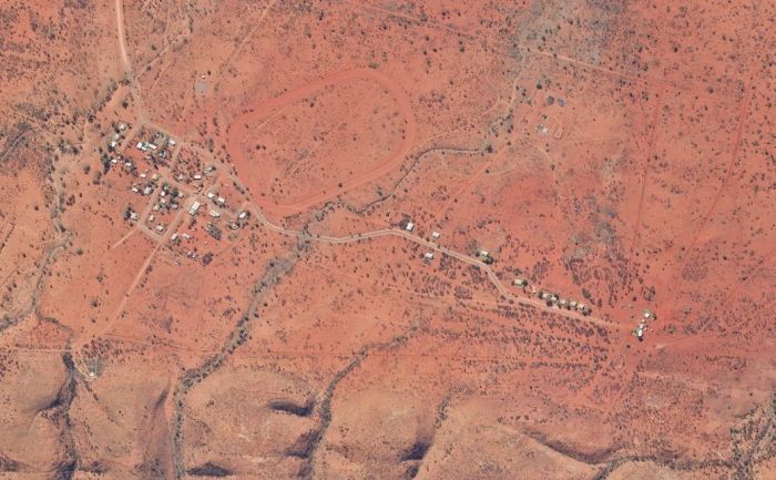 Aerial view of a small settlement in a reddish-brown arid landscape, with a few buildings and a road winding through.