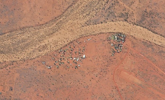 Aerial view of a small cluster of structures on red earth, near a dry riverbed with sparse vegetation.
