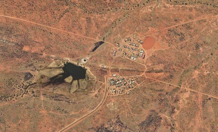 Overhead view of a small settlement in arid terrain, with buildings and roads. A pond or crater is visible on the left.