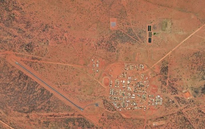 Aerial view of a small outback town in arid, reddish landscape, with an airstrip, buildings, and sports fields.
