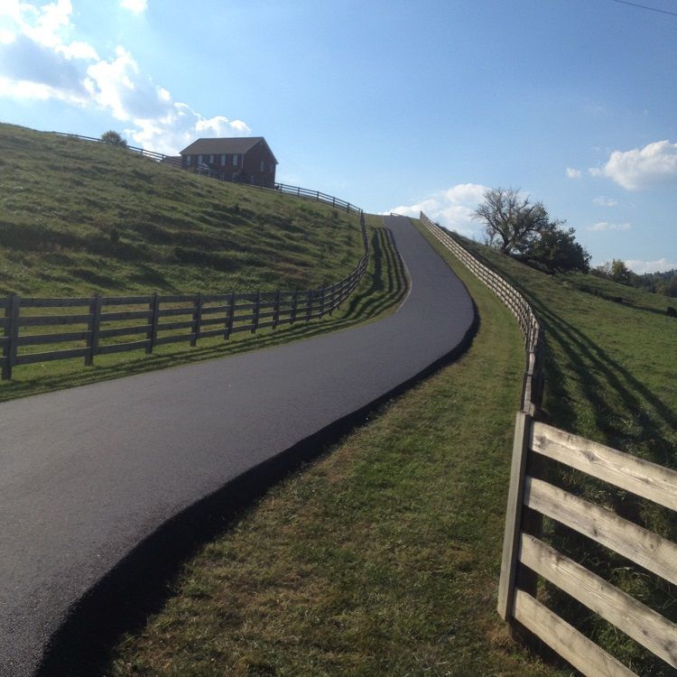 Asphalt driveway winds uphill past wooden fences, leading to a red-roofed house on a grassy hill.
