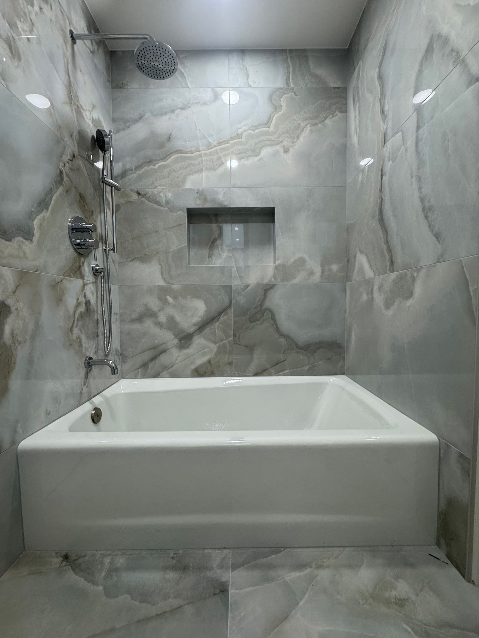 A white rectangular bathtub under a shower head, surrounded by grey and white veined marble tile walls with a small niche.