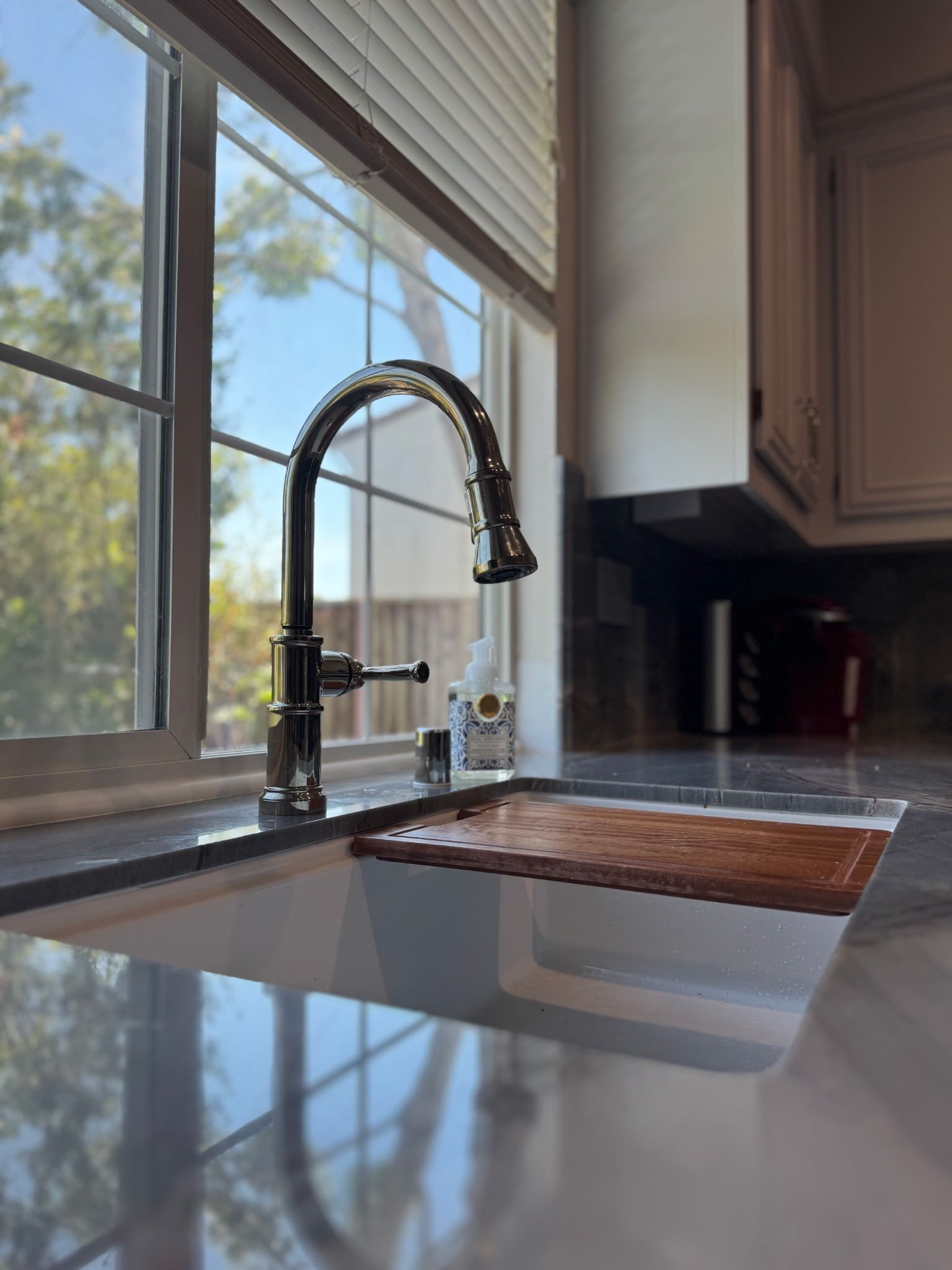 A chrome kitchen faucet sits above a white sink with a wooden drying mat, next to a window with white blinds.