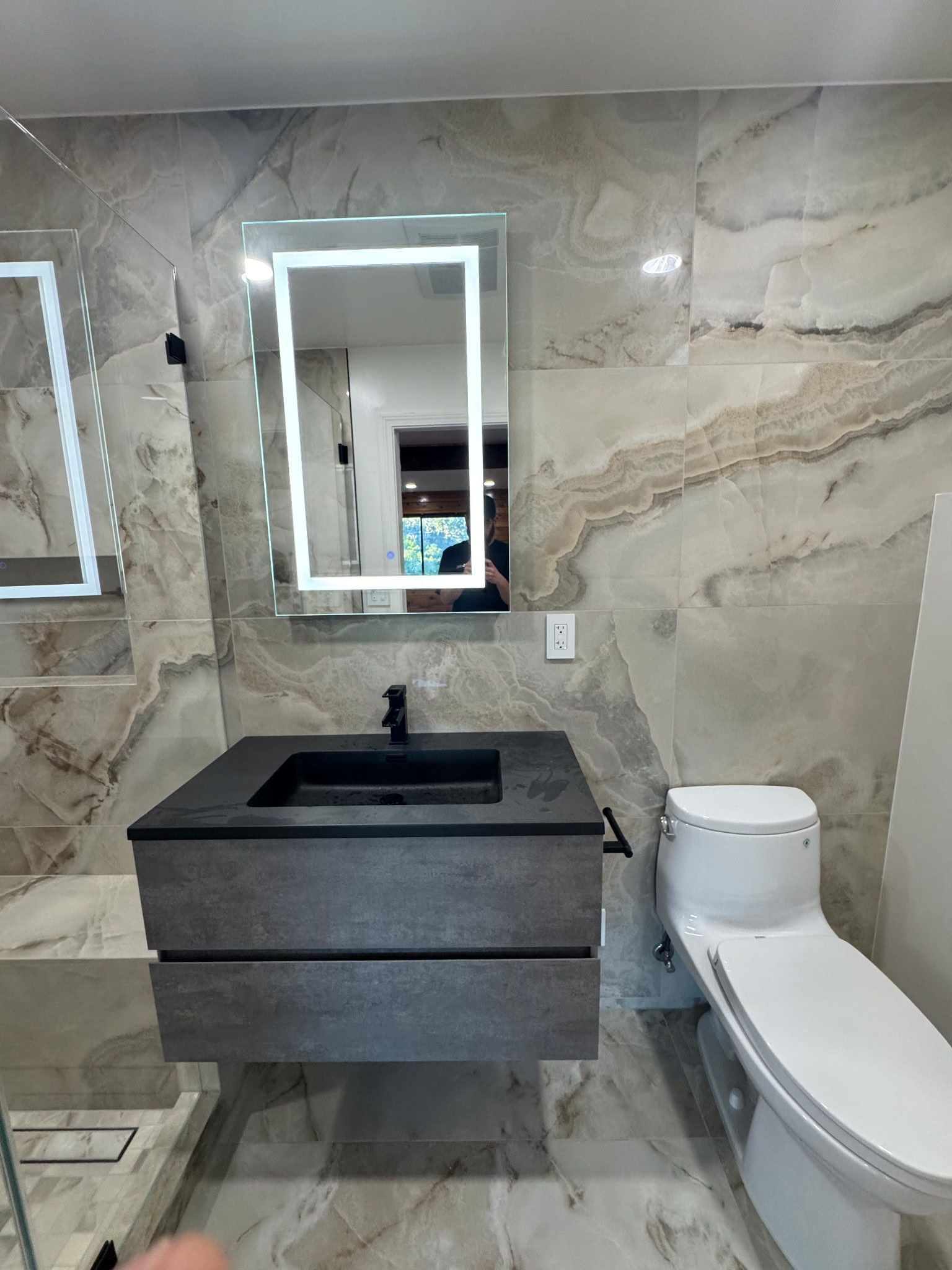 Modern bathroom with beige stone-patterned tiles, a floating dark wood vanity, black sink, and an illuminated mirror.
