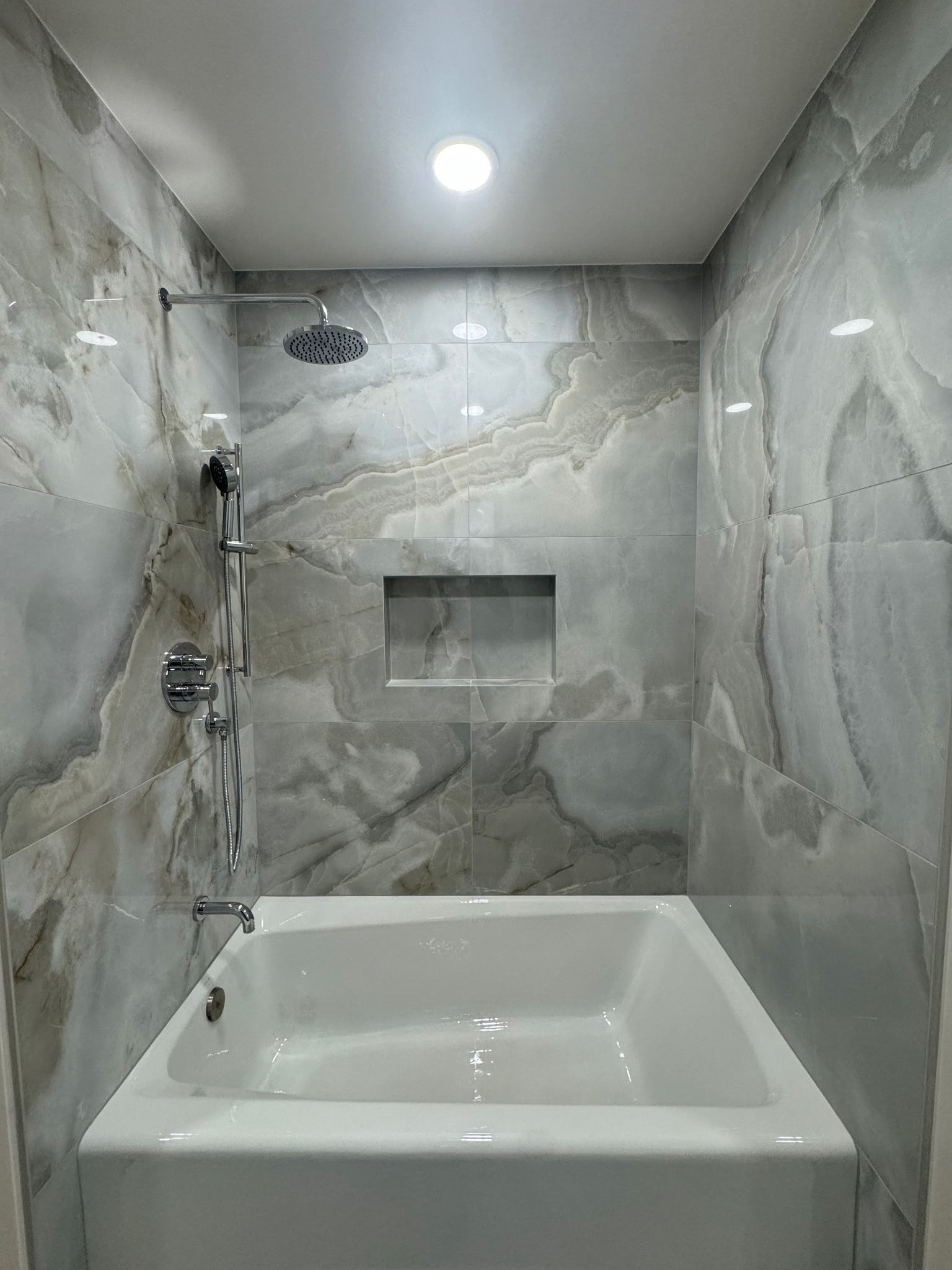 A white bathtub in a shower enclosure with light gray and tan marble-patterned tile walls and a recessed soap niche.