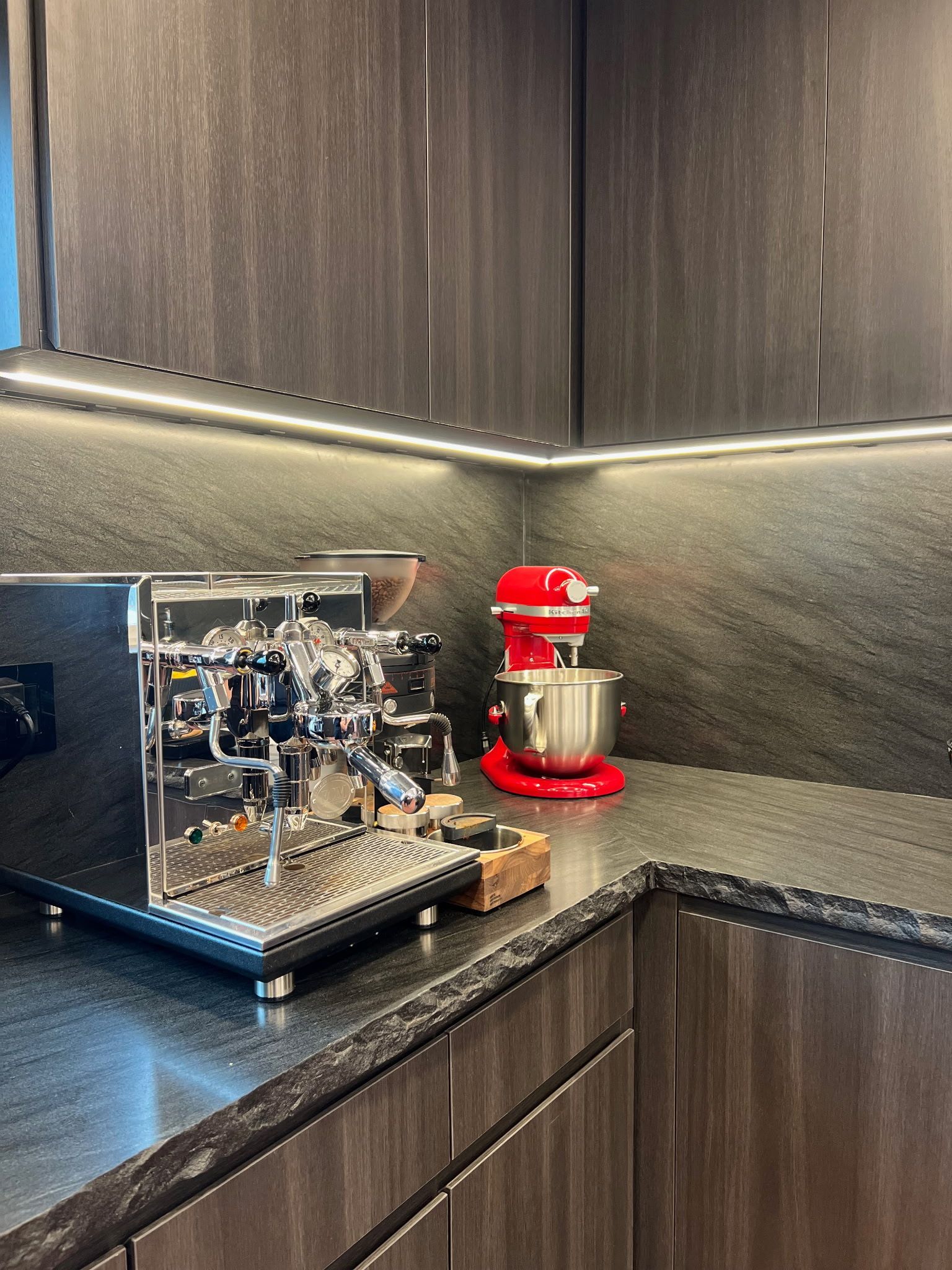A chrome espresso machine and a red stand mixer sit on a dark countertop in a kitchen with dark cabinetry and under-lighting.