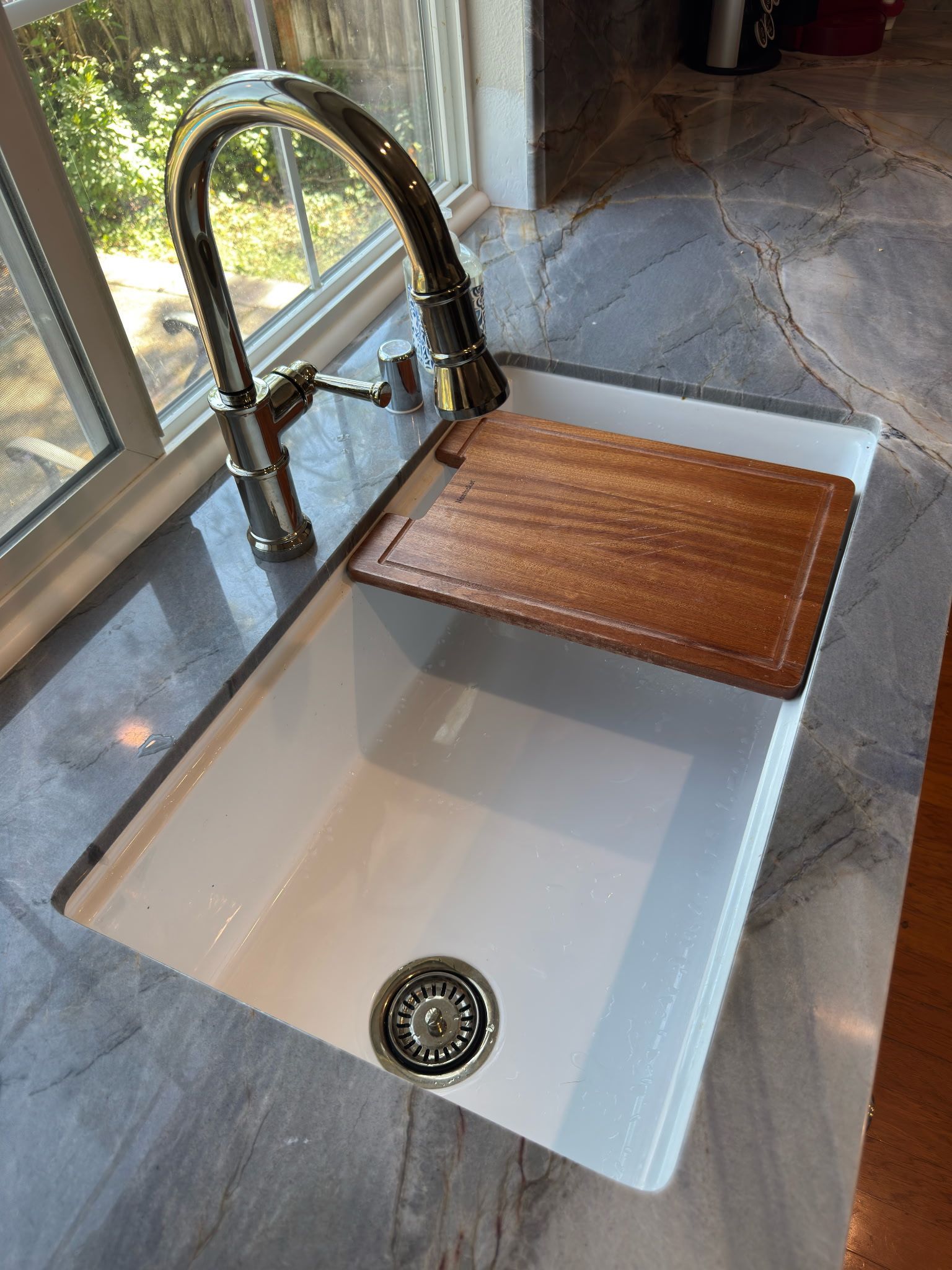 A white kitchen sink with a chrome faucet and a wooden cutting board nested over the basin, set in a marble countertop.