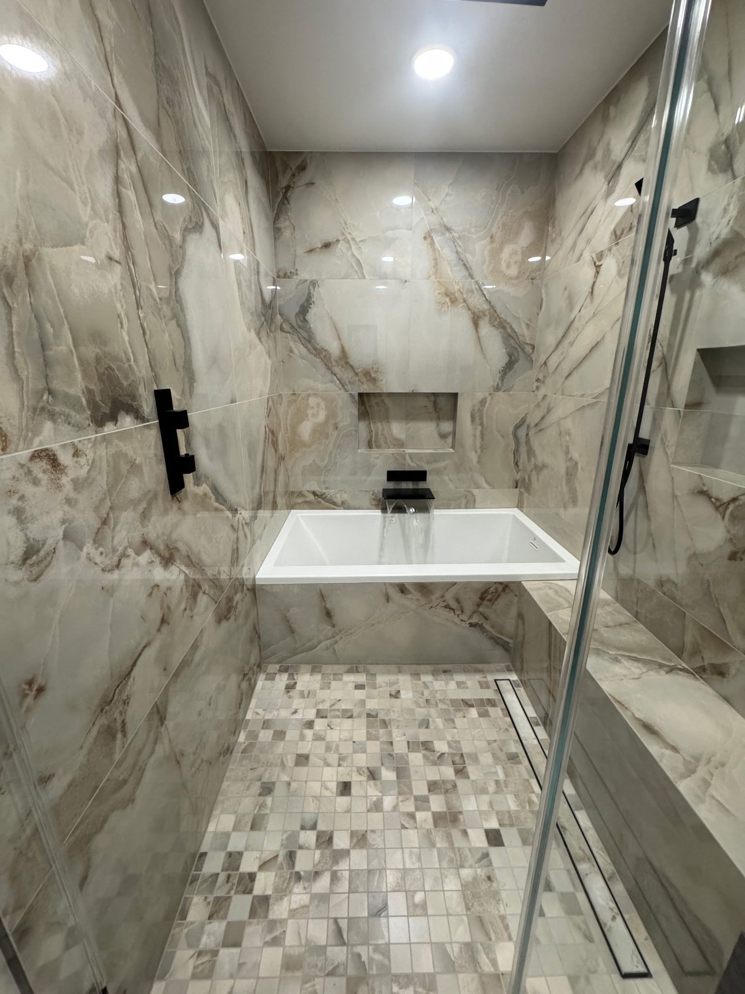 A modern, marble-tiled walk-in shower featuring a recessed niche, a white soaking tub, and a mosaic-tiled floor.