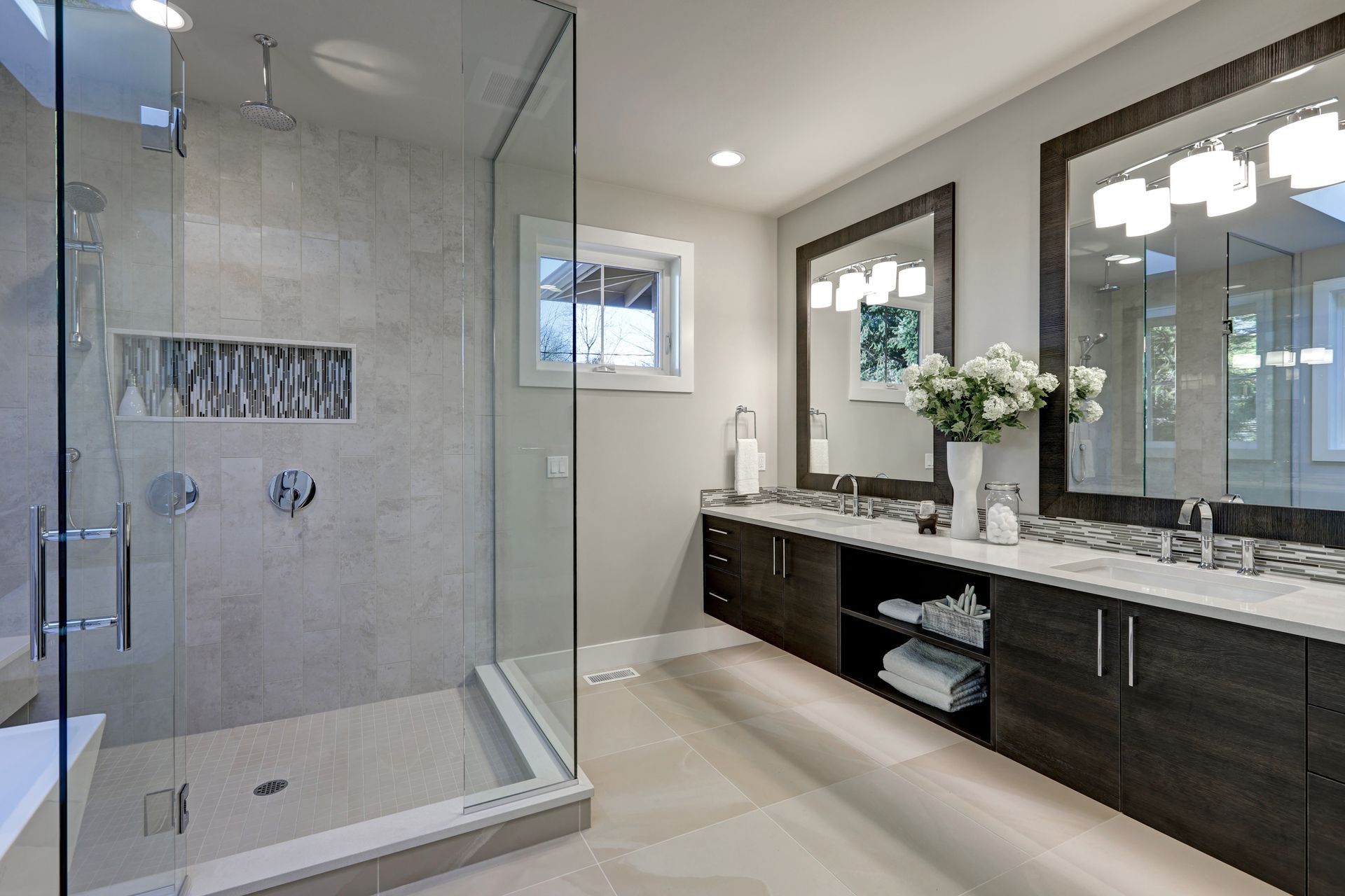 A modern bathroom featuring a glass-enclosed shower, dark wood vanity with dual sinks, two mirrors, and a bright window.