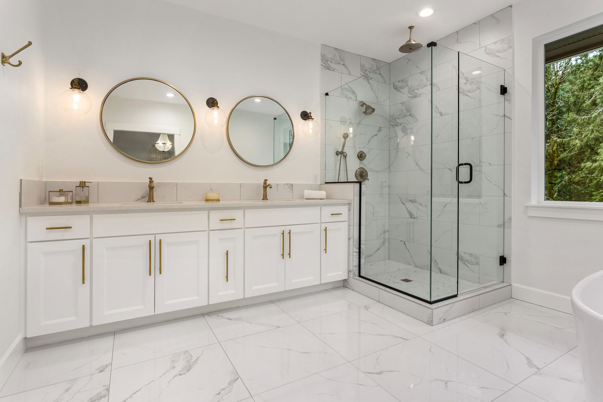 Modern bathroom with a double white vanity, gold hardware, two round mirrors, and a glass-enclosed marble walk-in shower.