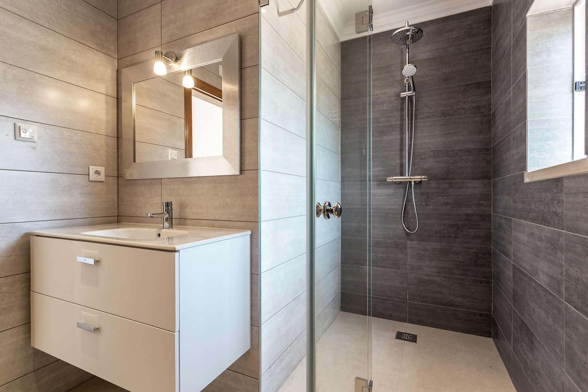 Modern bathroom with a white vanity, a silver-framed mirror, and a glass-enclosed shower with dark charcoal wall tiles.