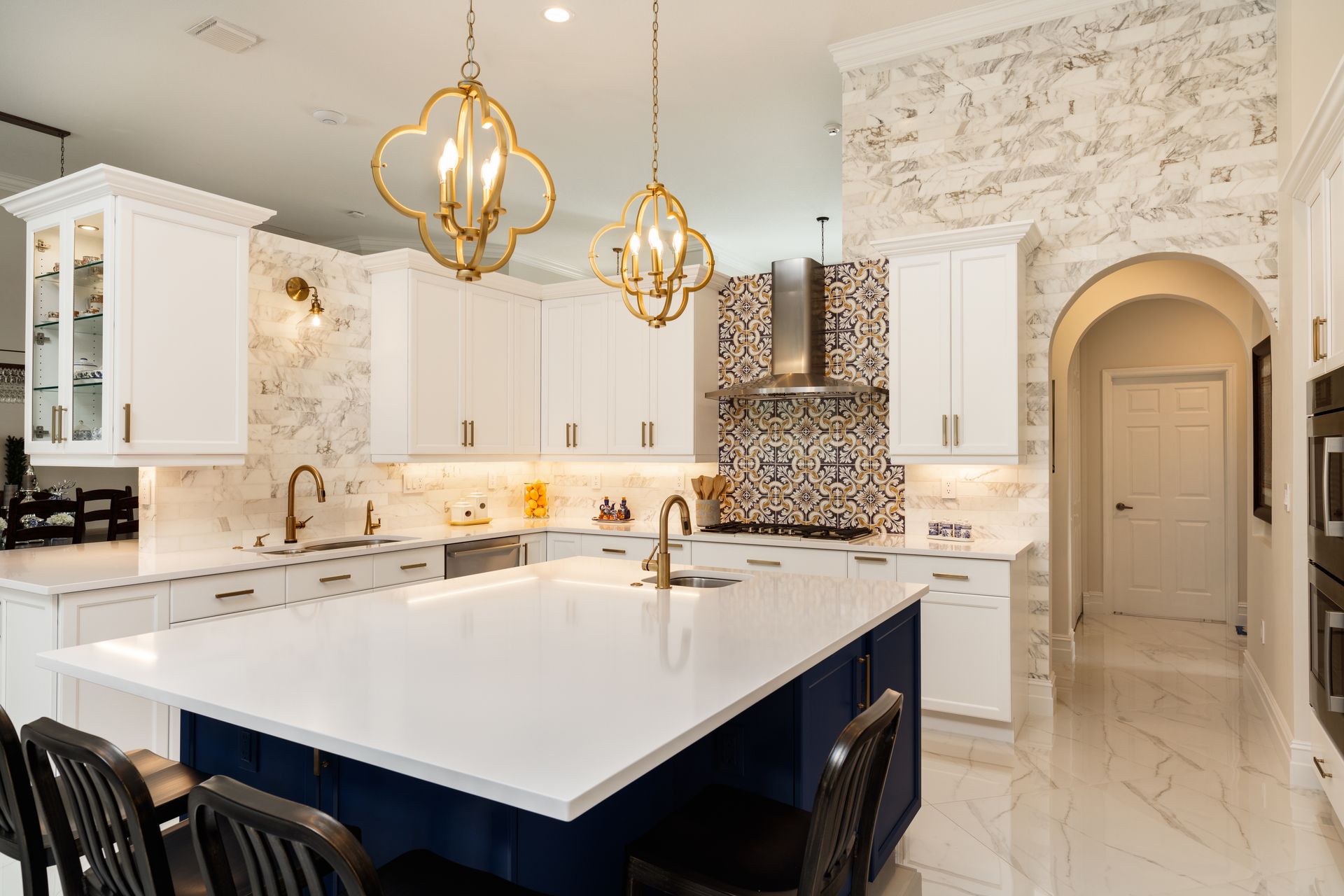 A bright kitchen featuring a large navy island with white countertops, gold pendant lights, and patterned tile backsplash.