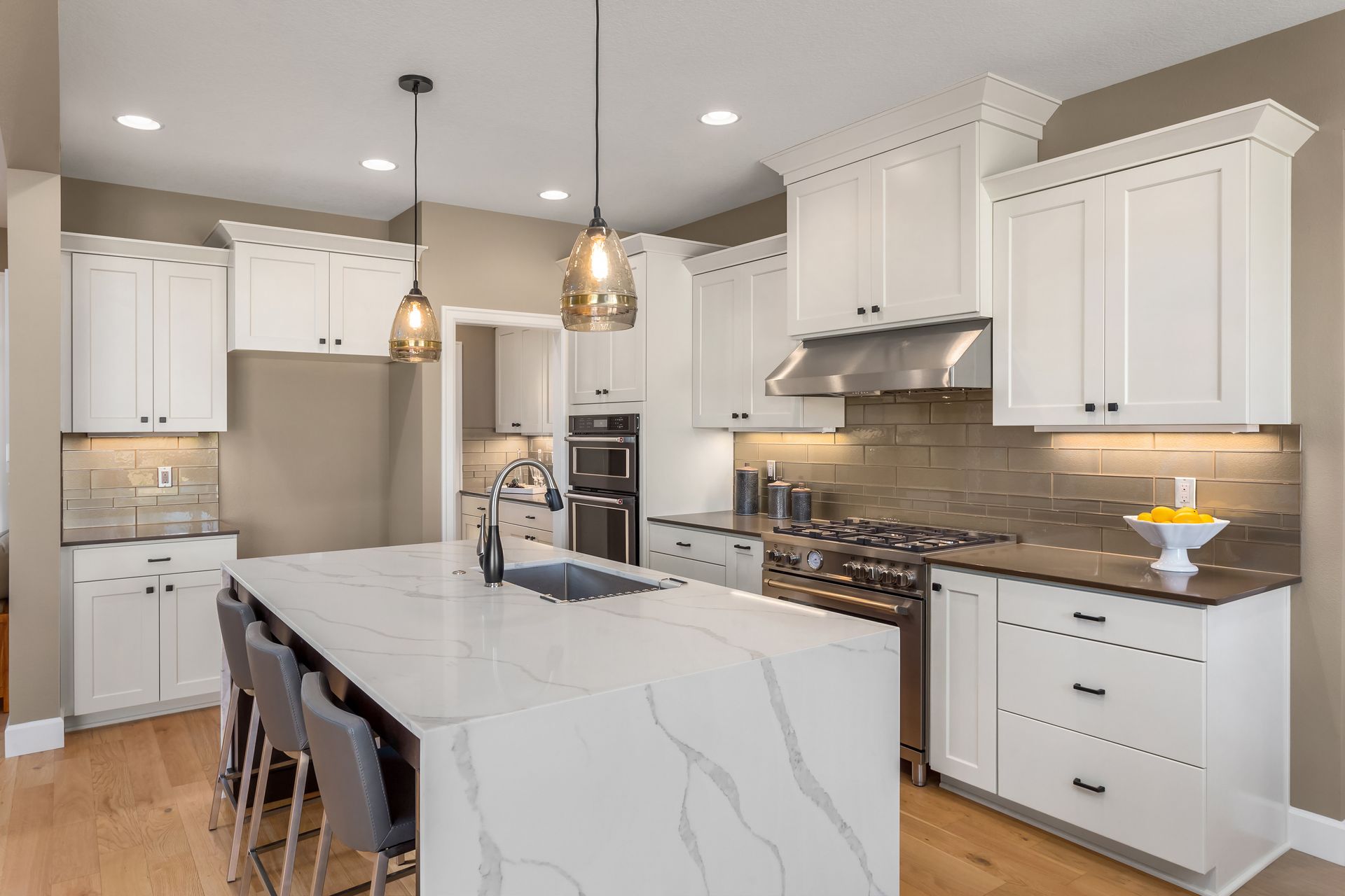 A modern kitchen with white cabinets, light wood floors, a large white marble island, and two pendant lights.