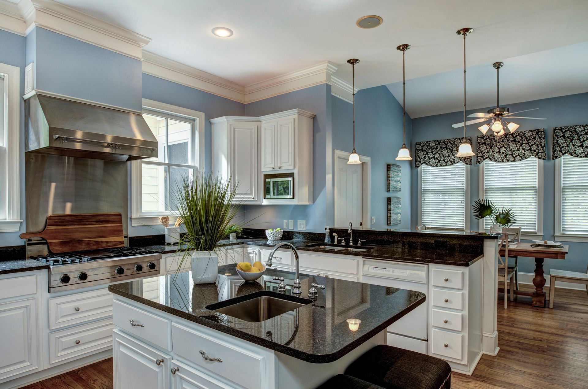 A bright kitchen with blue walls, white cabinets, a black granite island with a sink, and a stainless steel range hood.