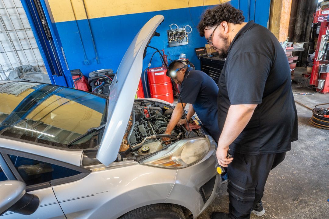 Two mechanics working on a car engine in a garage. One holds a tool, the other leans in close.