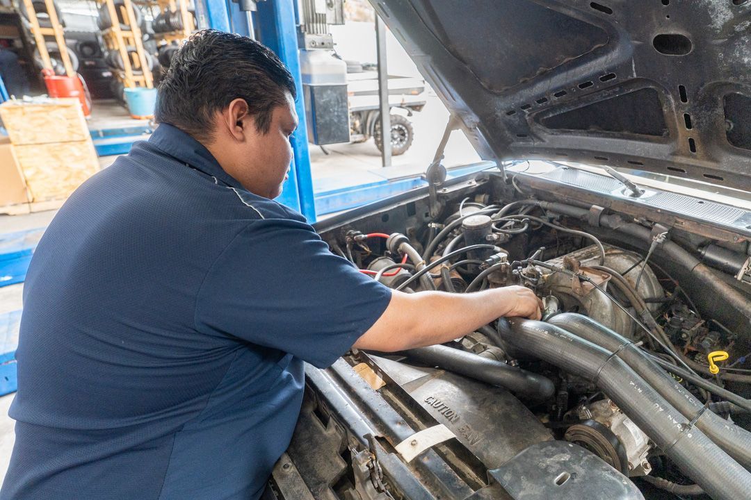 Mechanic working on the engine of a blue car with the hood open, inside a repair shop.