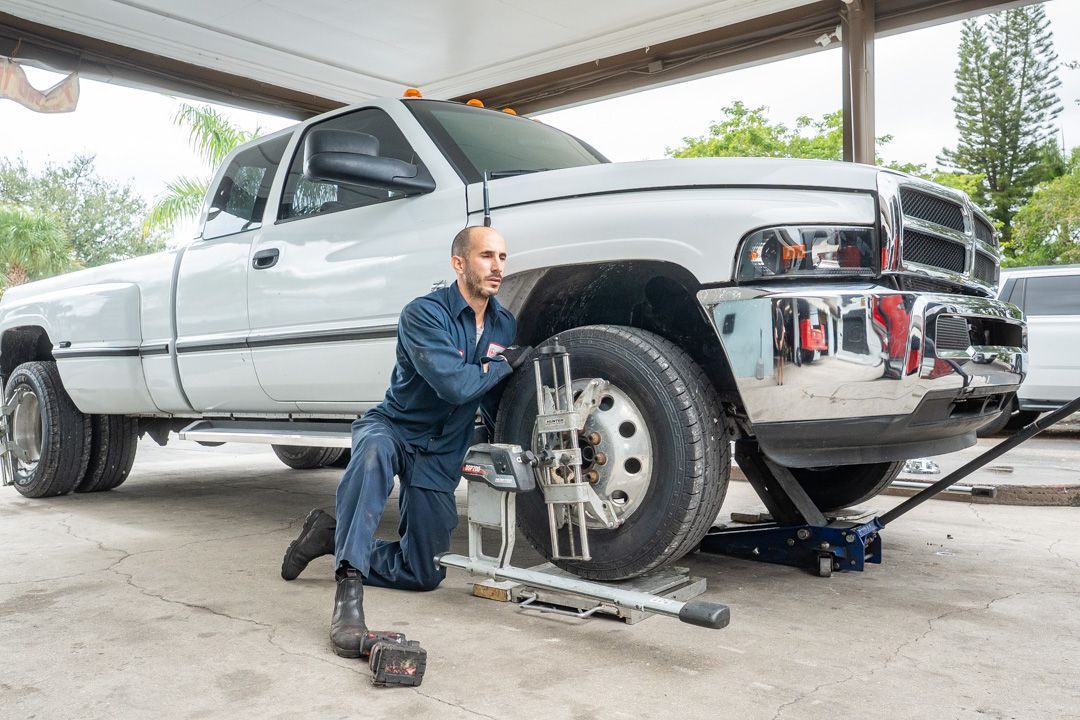 Mechanic adjusting truck wheel alignment in a shop. White truck, blue jumpsuit, tools visible.