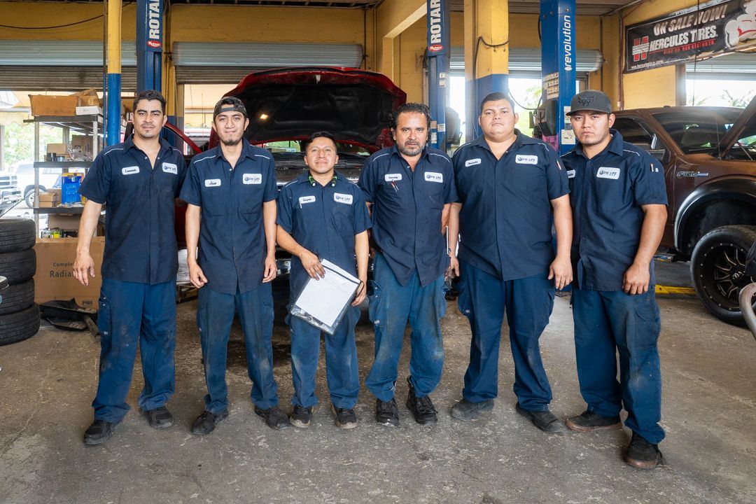 Six auto mechanics in blue uniforms pose in a garage with cars and equipment.