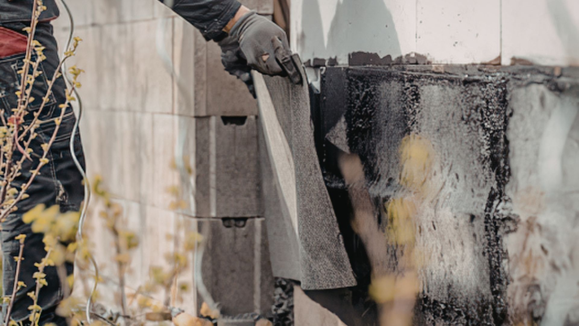 A person in work gloves peels a layer of black adhesive waterproofing membrane off a foundation wall.