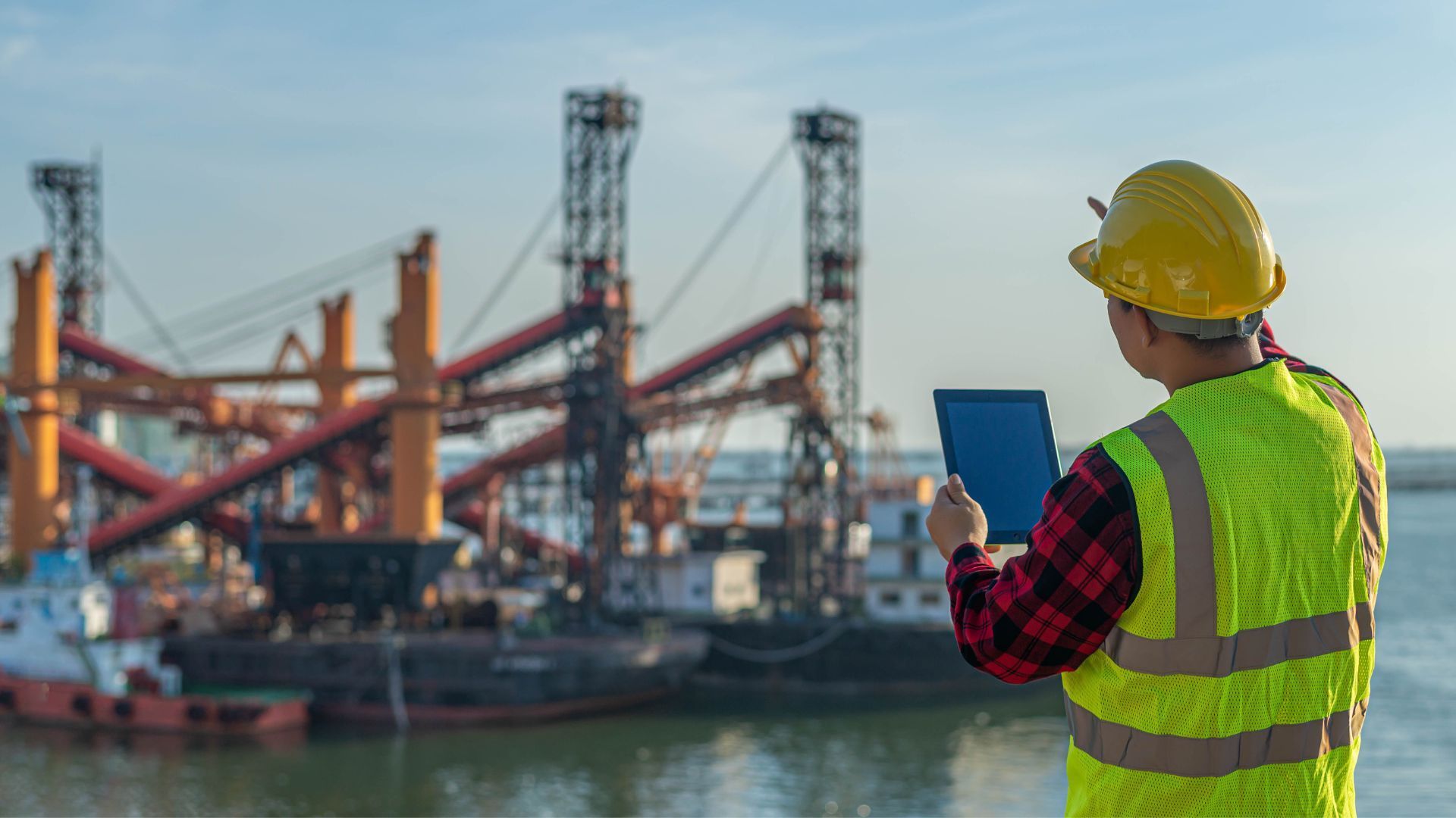 A construction worker in a hard hat and neon vest holds a tablet while viewing industrial cranes at a harbor.